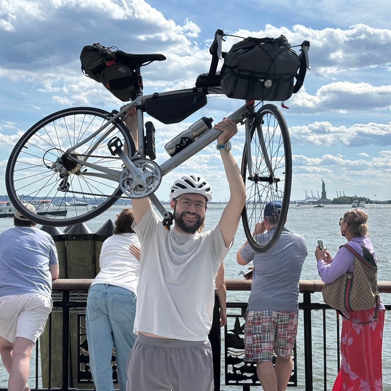 Graham Hagenah in a bike helmet, smiling and holding his silver road bike with its packed bags over his head at the railing in Battery Park, the harbor and the Statue of Liberty behind him under a cloudy blue sky