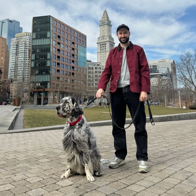 Graham Hagenah in a red jacket and cap, holding the leash of Philly, a black-and-white English setter sitting beside him, on the Rose Kennedy Greenway with Boston's Custom House tower behind them