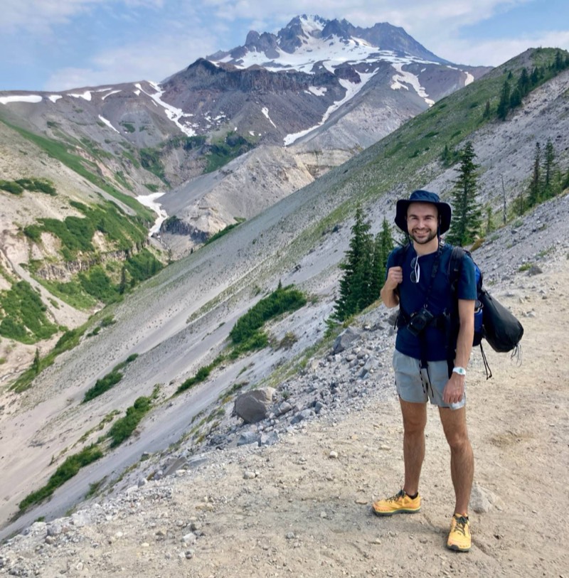 Graham Hagenah in a sun hat with a backpack and camera, smiling on a rocky trail, with the snowy peak of Mount Hood behind him