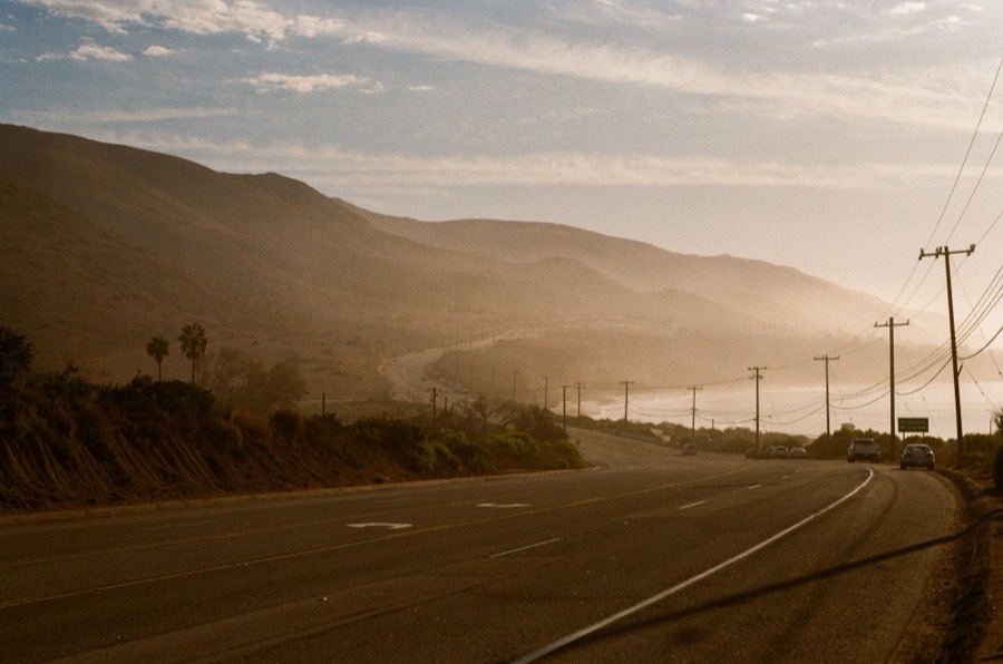 Photo by Graham Hagenah: a coastal highway in hazy golden light, telephone poles leading toward the ocean and layered hills