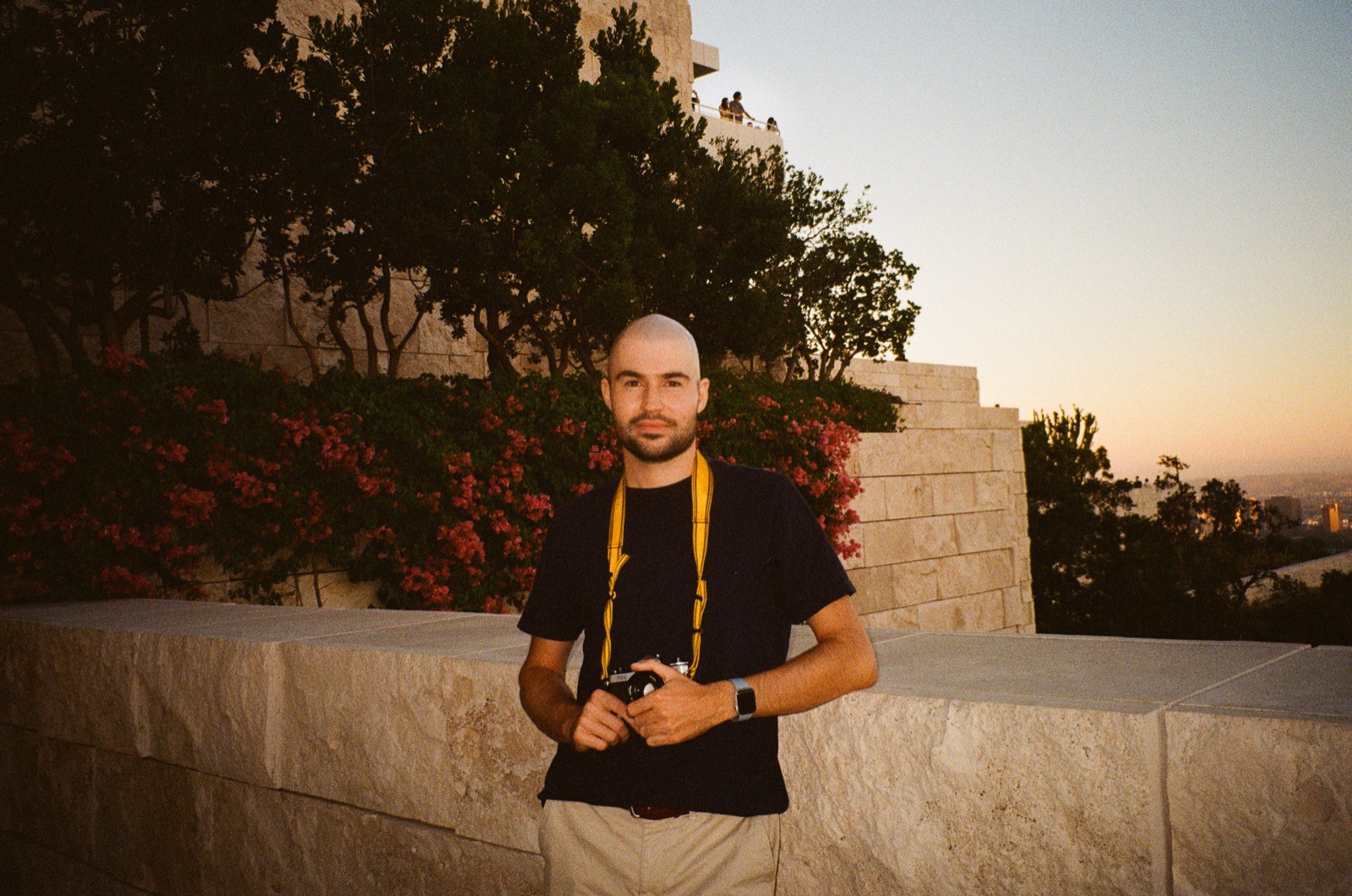 Graham at sunset at the Getty Center art museum in Los Angeles, holding a film camera on a yellow strap, in front of a stone wall and red flowering bushes, with the city skyline in the distance